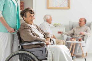Two older adults sit at a table painting colorful pictures together in a bright community room.